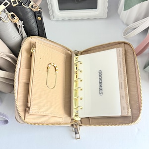 May include: Open, beige zippered planner with gold hardware. The planner is open to reveal a sectioned insert labeled "GROCERIES." A small gold chain with a charm is visible on the left side. The planner is on a white surface.