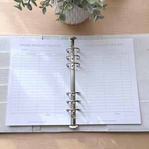 May include: A white, textured binder with a silver metal ring mechanism, holding a printed sheet labeled "ONLINE SPENDING TRACKER." The sheet has columns for date, description, category, and amount. A small plant in a white pot sits in the background.