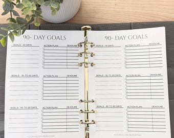 May include: Open binder with gold rings and pages titled "90-DAY GOALS." The pages are divided into sections for goals, action plans, and deadlines. A small plant in a white pot is visible in the background.