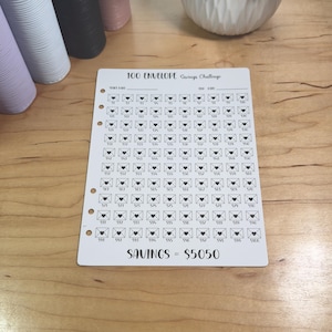 May include: A white savings challenge tracker with a grid of 100 envelope illustrations, each with a heart. The text "100 Envelope Savings Challenge" is at the top, and "SAVINGS = $5050" is at the bottom. The tracker is on a wooden surface.