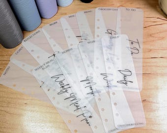 May include: A set of translucent plastic planner dividers with punched holes along the side. Each divider features printed black text labels including Weekly Meal Plan, Month, Week, Daily, To Do, Grocery List, Menu, Misc, Work, Reflections, Goals, and Notes.