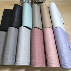 May include: A collection of ten different colored leather-bound journals. The journals are arranged in a row from left to right, with the colors ranging from black to light purple.