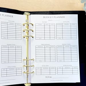 May include: A black binder with gold rings, open to a printed budget planner. The planner has sections for income, expenses, and savings. The words "Budget Planner" are at the top of the page. A plant is visible in the background.