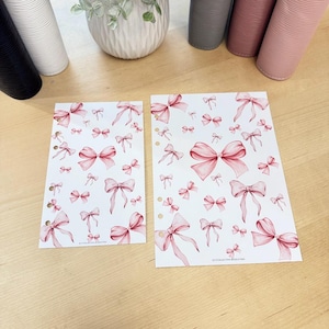 May include: Two white planner inserts with a repeating pattern of pink bows. The bows vary in size and are scattered across the pages. The pages have pre-punched holes for binding. The background is a light wood surface.