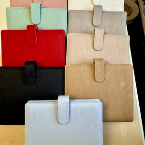May include: A stack of ten leather-bound journals in various colors, including pink, white, green, red, black, beige, and light blue. Each journal has a flap closure with a decorative strap.