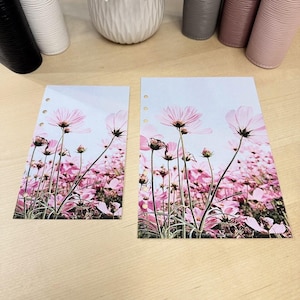 May include: Two planner inserts featuring a field of pink cosmos flowers against a light blue sky. The flowers are in full bloom, with delicate petals and green stems. The inserts have three holes for binding and are on a light wood surface.