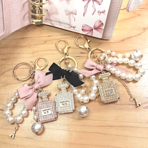 Puede incluir: Tres llaveros de frascos de perfume con herrajes dorados, detalles de perlas y lazos decorativos. Los frascos están adornados con pedrería y el texto "PARFUM N°5". Un llavero tiene un pequeño dije de la Torre Eiffel.