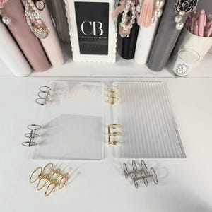 May include: Two rectangular acrylic binder covers, one clear and one ribbed, each featuring three-ring metal mechanisms. The set includes two additional loose three-ring metal mechanisms in gold and silver finishes, displayed on a white surface.