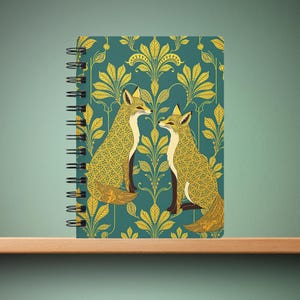 Può includere: Un quaderno a spirale blu-verde con una copertina a motivi dorati e gialli con due volpi che si guardano. Il motivo ricorda un design di carta da parati tradizionale.