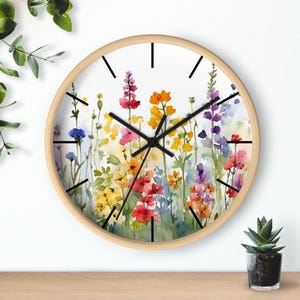 May include: A round wall clock with a light wood frame. The clock face features a watercolor painting of colorful wildflowers in shades of red, yellow, orange, purple, and blue. Black hands and hour markers complete the design.