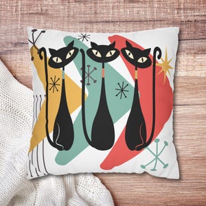 Könnte beinhalten: Ein weißes Kissen mit einem modernen Design aus der Mitte des Jahrhunderts, das drei schwarze Katzen mit langen Schwänzen und farbenfrohen abstrakten Formen im Hintergrund zeigt.