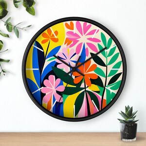 May include: A round wall clock with a black frame and a colorful floral design. The clock face features a yellow background with pink, orange, blue, and green flowers and leaves.