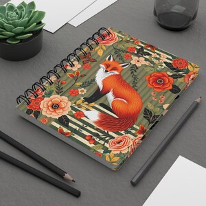 Può includere: Quaderno a spirale con un'illustrazione di una volpe con pelliccia arancione e petto bianco, circondata da fiori e strisce. Il design include rose, farfalle e un motivo a righe verdi e bianche. Un accessorio di cancelleria decorativo.
