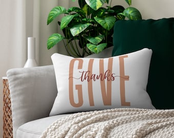 May include: White throw pillow with the text "Give thanks" in a brown color. The pillow is on a couch with a white blanket and a green pillow.
