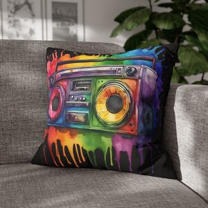 Puede incluir: Una almohada negra con una ilustración acuarela colorida de una radiocassette. La radiocassette está rodeada de salpicaduras de pintura de colores arcoíris.