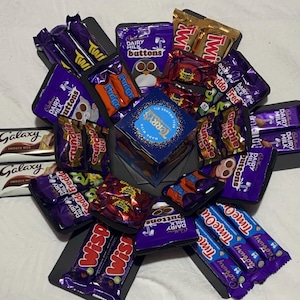 May include: A selection of wrapped chocolate bars and sweets, such as Galaxy, Twix, and Cadbury Dairy Milk Buttons, presented in a decorative box. The box includes a central blue box with the text "Terry's".