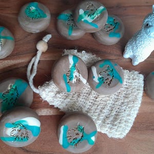 May include: A collection of round, brown soaps with teal and white accents, featuring a wolf design. A white, textured loofah and a white llama figurine are also present. The soaps are arranged on a wooden surface.