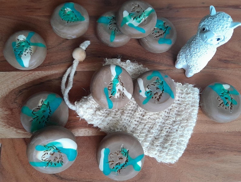 May include: A collection of round, brown soaps with teal and white accents, featuring a wolf design. A white, textured loofah and a white llama figurine are also present. The soaps are arranged on a wooden surface.