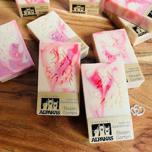 May include: Artisan soap bars with a cream base and pink marbling. Each bar features a carved alpaca design and a label with the text "ALPAKAS Garten". The soaps are arranged on a wooden surface.