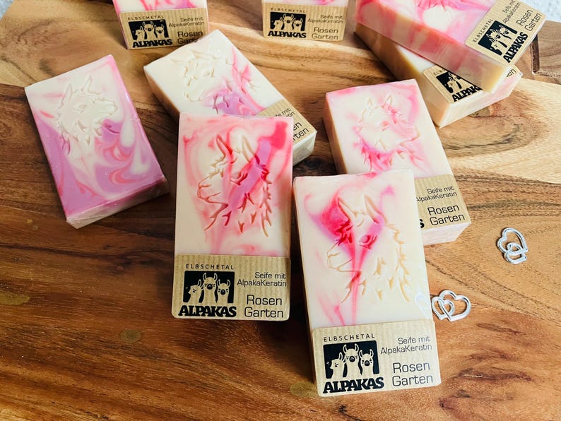 May include: Artisan soap bars with a cream base and pink marbling. Each bar features a carved alpaca design and a label with the text "ALPAKAS Garten". The soaps are arranged on a wooden surface.