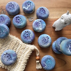 May include: A collection of round, marbled blue soaps with a wolf design, alongside a small white llama figurine and a natural fiber washcloth. The soaps have a swirling pattern of blue and purple. The background is a wooden surface.