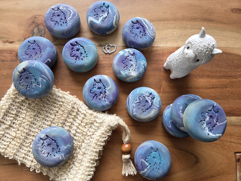 May include: A collection of round, marbled blue soaps with a wolf design, alongside a small white llama figurine and a natural fiber washcloth. The soaps have a swirling pattern of blue and purple. The background is a wooden surface.