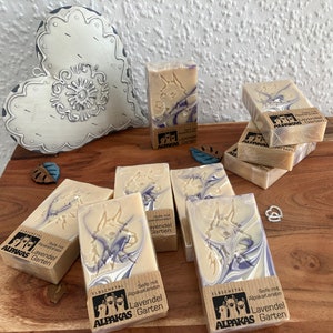 May include: Several bars of soap with a light beige base and purple swirls are arranged on a wooden surface. Each bar is wrapped and features a carved design and the text "ALPAKAS Garten". A decorative heart is in the background.