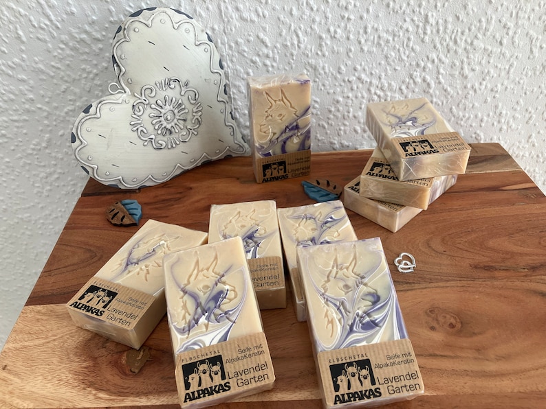 May include: Several bars of soap with a light beige base and purple swirls are arranged on a wooden surface. Each bar is wrapped and features a carved design and the text "ALPAKAS Garten". A decorative heart is in the background.