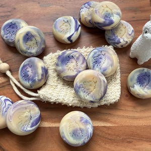 May include: A collection of round, marbled soaps in shades of purple, cream, and yellow, each with a wolf design. A white ceramic alpaca figurine and a natural fiber soap bag are also present on a wooden surface.