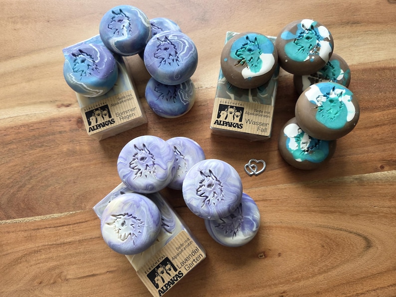 May include: Artisan soaps with alpaca designs in various colors, including blue, purple, and brown. The soaps are round and feature an alpaca silhouette. Some soaps are paired with small, rectangular boxes with text.