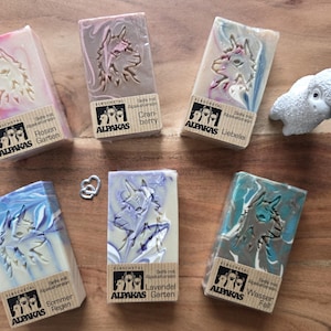 May include: A collection of artisan soaps with alpaca designs and various colors, including pink, blue, and teal. Each soap bar has a unique swirl pattern and is labeled with German text. A small white alpaca figurine is also present.