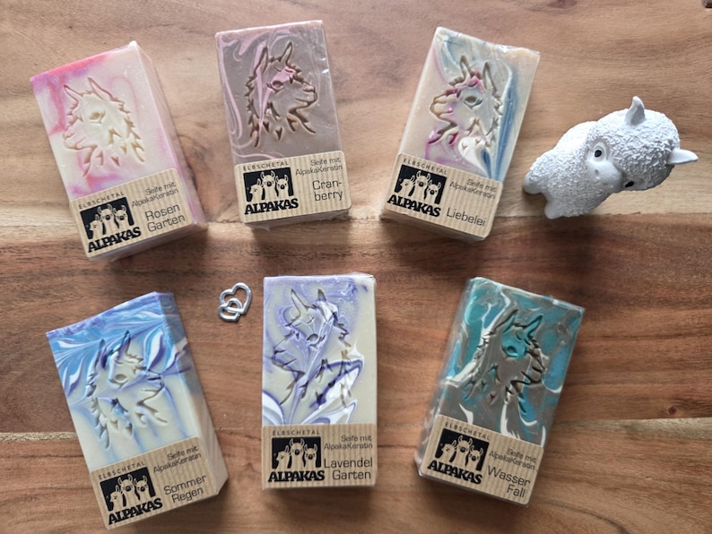 May include: A collection of artisan soaps with alpaca designs and various colors, including pink, blue, and teal. Each soap bar has a unique swirl pattern and is labeled with German text. A small white alpaca figurine is also present.