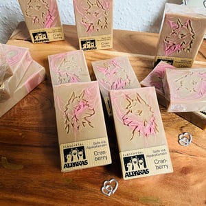 May include: Artisan soaps with a light brown base and pink accents, featuring an embossed llama design. The soaps are labeled with the brand "ALPAKAS" and "Cranberry". Several bars are arranged on a wooden surface.