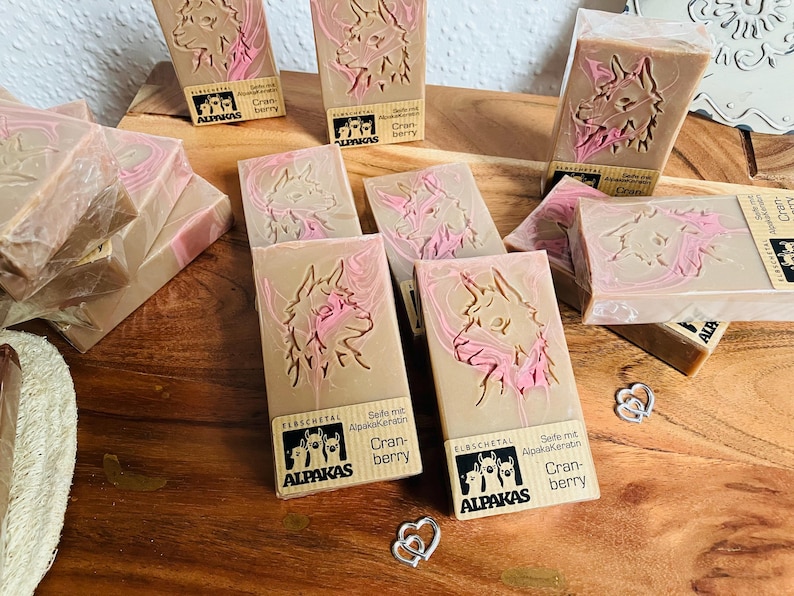 May include: Artisan soaps with a light brown base and pink accents, featuring an embossed llama design. The soaps are labeled with the brand "ALPAKAS" and "Cranberry". Several bars are arranged on a wooden surface.