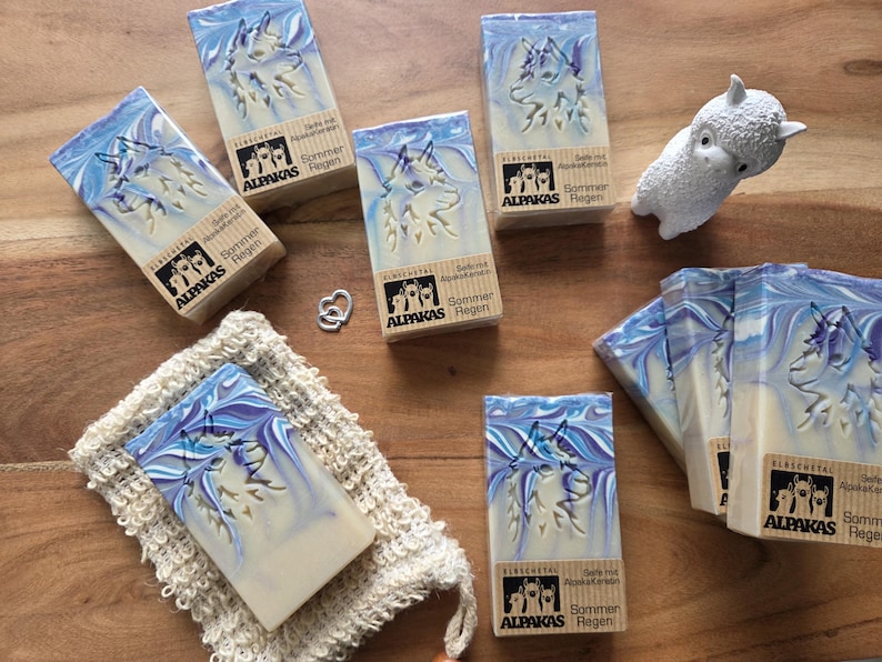 May include: Several bars of soap with a blue and white swirl design and a wolf imprint. The soap bars are packaged in boxes with the text "ALPAKAS Regen" and "Sommer". A small white alpaca figurine and a washcloth are also present.
