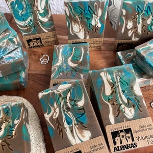 May include: Artisan soap bars with a marbled design in shades of blue, brown, and white. Each bar features a carved design and a small label with text. The soaps are arranged on a wooden surface.
