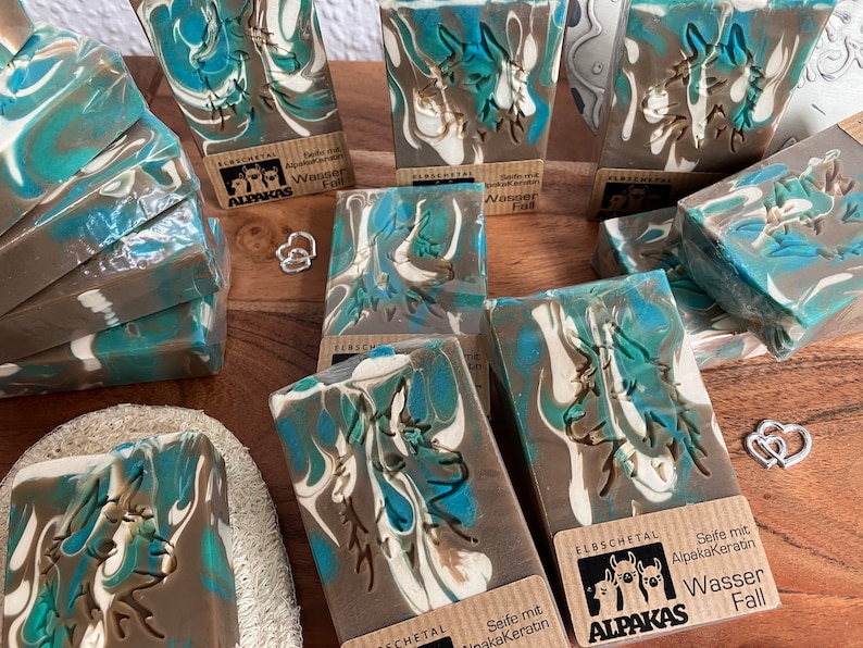 May include: Artisan soap bars with a marbled design in shades of blue, brown, and white. Each bar features a carved design and a small label with text. The soaps are arranged on a wooden surface.
