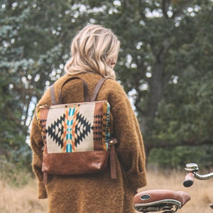 May include: A brown and tan backpack with a geometric pattern is worn by a person in a brown coat. A black bicycle with a brown leather seat and a wicker basket is in the foreground. The scene is set outdoors with tall grass and trees.