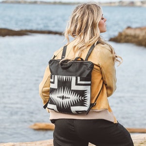 Pode incluir: Uma mulher vestindo uma jaqueta de camurça marrom e calças pretas está em pé em um tronco. Ela está usando uma mochila preta com um padrão geométrico branco e preto.