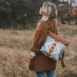 May include: A woman wearing a brown sweater and blue jeans is standing in a field of tall grass. She is carrying a blue and brown woven backpack with leather straps.