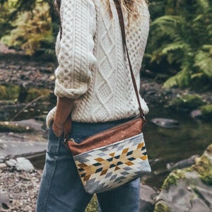 May include: A cream-colored cable knit sweater and blue jeans. A brown leather strap crossbody bag with a geometric pattern in shades of gray, yellow, and brown is worn over the shoulder. The background is a forest with a stream.