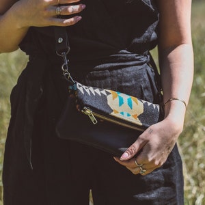 May include: Black leather clutch with a patterned fabric flap featuring geometric shapes in teal, yellow, and white. The clutch has a gold zipper and a black leather strap. The person is wearing a black jumpsuit.