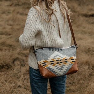 Puede incluir: Una mujer con un su&eacute;ter de punto blanco y jeans azules lleva un bolso bandolera de estampado gris y amarillo con una solapa de cuero marr&oacute;n.
