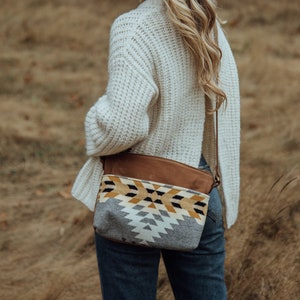 May include: A crossbody bag with a brown leather top and a patterned bottom in grey, yellow, and black geometric shapes. The bag is worn over a white sweater and blue jeans.
