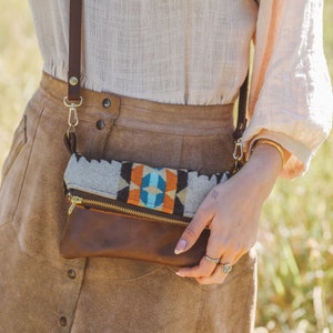 Peut inclure: Un sac bandoulière en cuir marron avec un rabat en tissu à motifs. Le sac est doté d'une fermeture éclair dorée et d'une bandoulière en cuir marron. Le rabat en tissu présente un motif géométrique dans les tons de bleu, orange et gris.