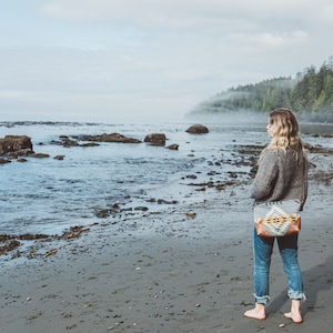 Puede incluir: Una mujer con un su&eacute;ter gris y jeans azules se para en una playa de arena mirando hacia el oc&eacute;ano. Lleva un bolso tejido de colores con una correa de cuero marr&oacute;n.
