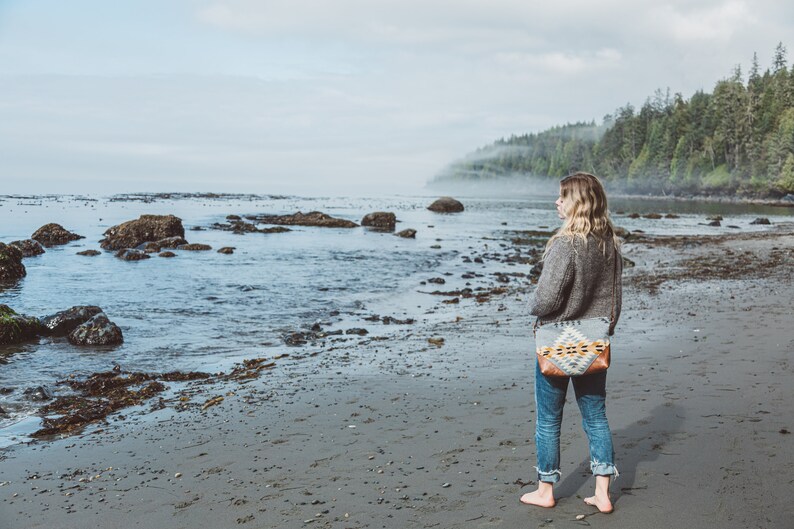 Puede incluir: Una mujer con un su&eacute;ter gris y jeans azules se para en una playa de arena mirando hacia el oc&eacute;ano. Lleva un bolso tejido de colores con una correa de cuero marr&oacute;n.