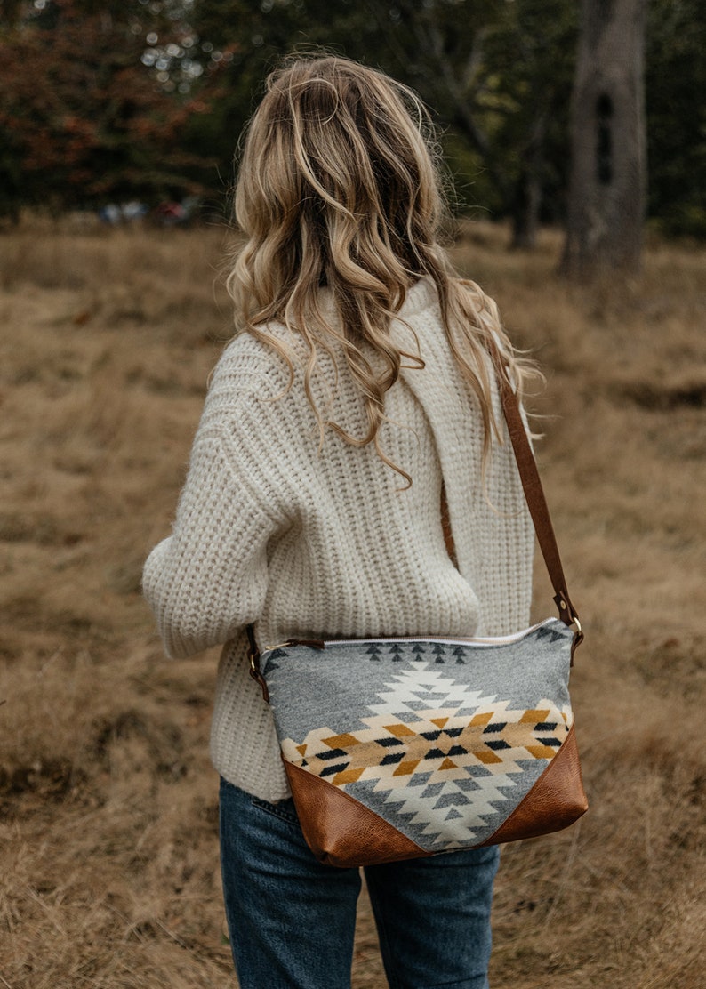 Puede incluir: Una mujer con un su&eacute;ter de punto blanco y jeans azules lleva un bolso bandolera de estampado gris y amarillo con una solapa de cuero marr&oacute;n.