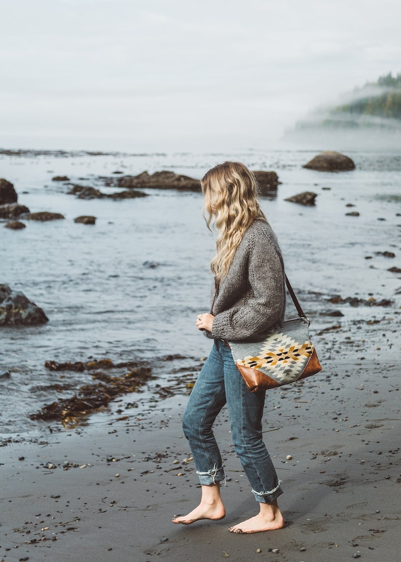 Puede incluir: Una mujer camina descalza por una playa de arena, con un c&aacute;rdigan de punto gris y unos vaqueros azules. Lleva un bolso tejido con un patr&oacute;n geom&eacute;trico y detalles de cuero marr&oacute;n.