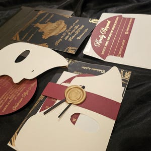 Pode incluir: Uma variedade de convites de casamento e cartões RSVP. Os convites apresentam um design preto e dourado com um recorte em forma de máscara. Os cartões RSVP são bordô e dourados. Um selo de cera dourada é visível.